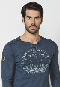 Dlouhá rukávová navy blue henley košile z texturované tkaniny. Obsahuje grafický potisk s motivem motocyklů a knoflíkovou légou.