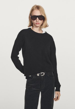 Sort strikket sweater med rund hals, båret over en hvid skjorte. Matchende med sorte corduroy-bukser og mørke solbriller.