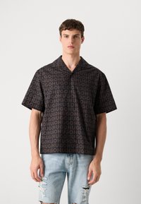 Camisa negra de manga corta con un patrón de logotipo marrón repetido, con collar y cierre de botón, combinada con jeans de mezclilla claros y desgastados.