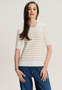 Mujer con cabello largo castaño, llevando pendientes de aro dorados, un suéter de manga corta de canalé blanco con rayas beiges y jeans azules, de pie contra un fondo simple.
