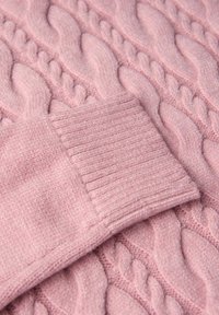 Weiches, rosa gestricktes Material mit einem gerippten Bündchen-Detail vor einem Kabelstrickmuster-Hintergrund.