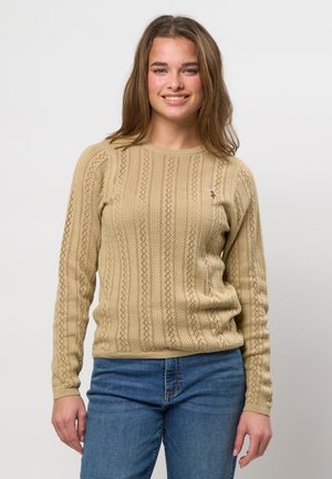 Młoda kobieta uśmiechająca się, ubrana w beżowy sweter w wzory i niebieskie dżinsy, stojąca na tle jednolitego białego tła.