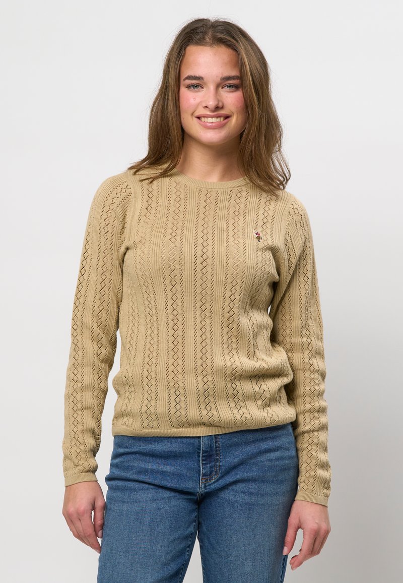 Jeune femme souriante, portant un pull en tricot beige à motifs et un jean bleu, debout devant un fond blanc uni.