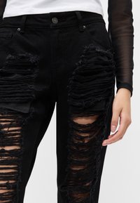 Pantalones de mezclilla negra con textura desgastada, que presentan grandes áreas rasgadas y bordes deshilachados. Diseño clásico de botón y cinco bolsillos.