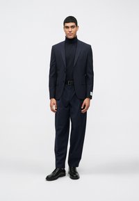 Μαρκάζι ναυτικού με κομψό σακάκι και παντελόνι με φαρδιά πόδια, σε συνδυασμό με εφαρμοστό πουλόβερ με ψηλό λαιμό. Λείος υφασμάτινος συνδυασμός, μαύρα δερμάτινα παπούτσια, λεπτομέρειες με διακριτική φινέτσα.