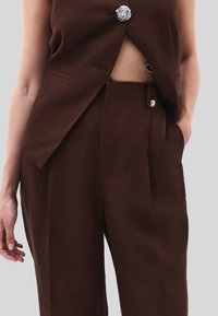 Hnědá lněná vesta s otevřenou stranou, upevněná dekorativními stříbrnými knoflíky. Odpovídající kalhoty mají boční kapsy a podobné detaily s knoflíky.