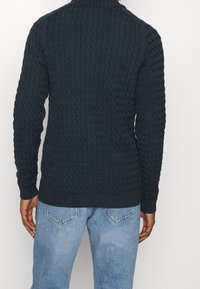 Man som bär en mörk marinblå strukturerad stickad tröja och ljusblå jeans, sedd bakifrån mot en enkel vit bakgrund.