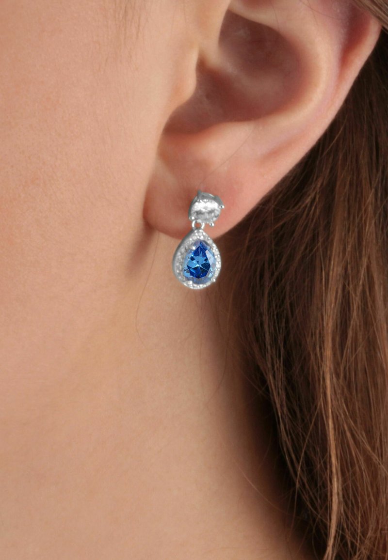 Gli orecchini a goccia presentano una gemma blu con faccette, circondata da pietre trasparenti, montata in metallo tono argento. Chiusura a bottone.