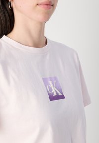 T-shirt in cotone rosa chiaro con una grafica rettangolare viola al centro e il logo bianco "cK". Girocollo e maniche corte.