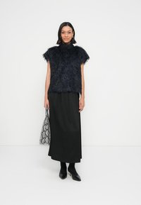 Černý vršek s chlupatou texturou, krátké rukávy, vysoký výstřih, zkombinovaný s dlouhou černou sukní a botami na podpatku, držící kabelku se vzorem hadí kůže.