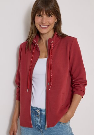Kvinde smiler iført en rød lynlåstrøje med snore over en hvid top og blå jeans, stående mod en ensartet lys baggrund.
