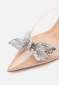 Chaussure à talon haut nude pointue avec côtés transparents et grande décoration nœud en argent ornée de bijoux à l'avant.