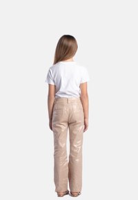 Pantaloni flare lucidi di un beige chiaro con vita alta, tasche posteriori e una superficie liscia abbinati a una maglietta bianca. Vista da dietro.