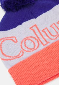 Adīts beanie ar purpura augšpusi, baltu vidu, kurā oranžā krāsā ir uzraksts "Columbia", un korallkrāsas ribota aproce. Pabeigts ar pomponu virsū.