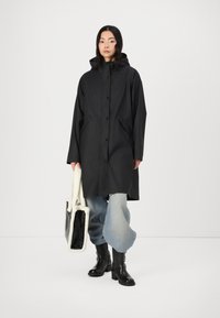 Svart lång kappa med huva, tryckknappar, sidofickor; kombinerad med ljusblå vida jeans och svarta klackstövlar. Bär en väskan fodrad med päls.