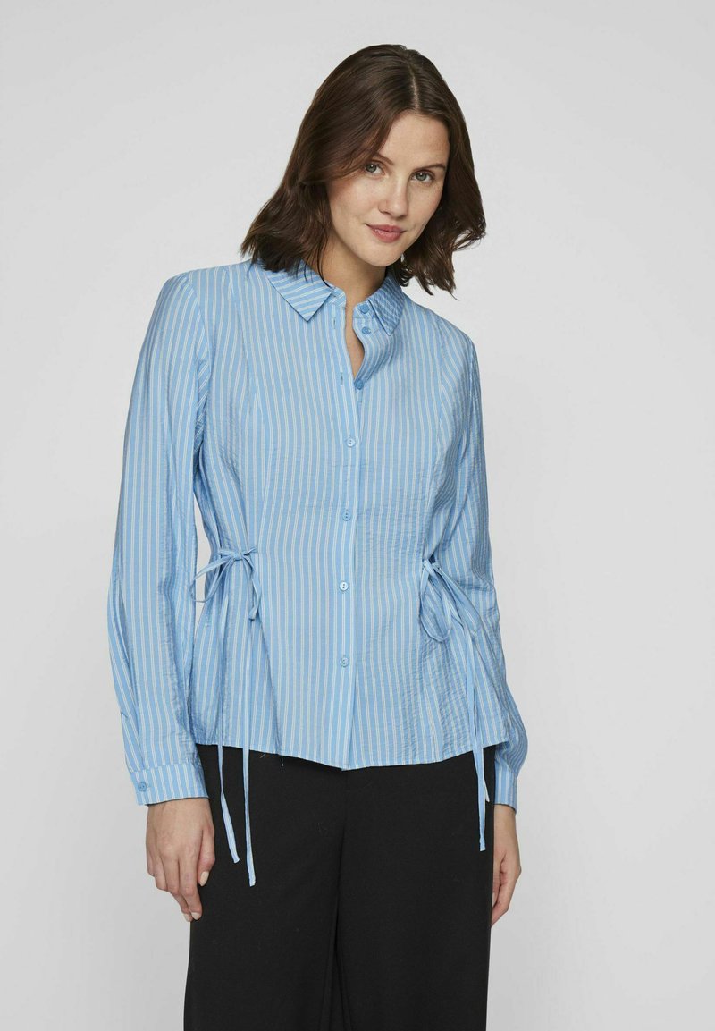 Camisa de botones a rayas azul claro con mangas largas, que presenta detalles de lazo en los lados y un dobladillo recto. Fabricada en una suave tela texturizada.