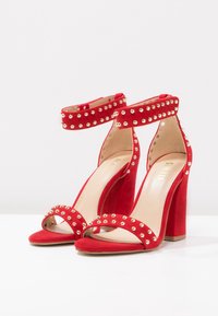 Sandalias de tacón alto de ante rojo con acentos de tachuelas doradas, tiras en el tobillo y diseño de punta abierta. Tacón grueso para mayor estabilidad. Forro interior suave.