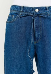 Pantaloni din denim de culoare bleu mediu, cu talie medie, având o curea legată, buzunare frontale și detalii de cusătură portocalie contrastantă.