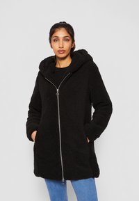 Manteau à capuche noir et fluffy avec fermeture éclair, doté d'une texture extérieure et de deux poches avant, associé à un jean bleu clair.