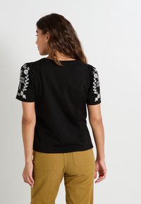 Top negro de manga corta con patrones florales bordados en los hombros, combinado con pantalones beige. Presenta un corte clásico y cuello redondo.