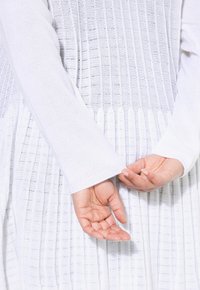 Vitt stickat plagg med ribbat mönster, långa ärmar och en plisserad design vid midjan. Händerna är placerade bakom ryggen.
