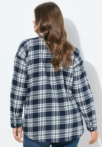 Frau mit langen braunen Haaren, die ein kariertes Button-Up-Hemd in Marineblau und Weiß trägt, steht mit dem Rücken zur Kamera.