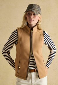 Beige Fleece-Weste mit hohem Kragen und goldenem Reißverschluss, über einem navy- und cremegestreiften Hemd getragen. Braune Akzente und Taschen. Gemusterte Mütze.