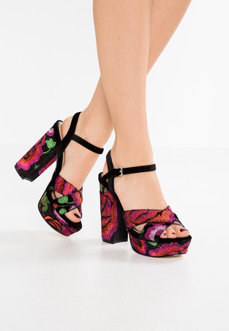 Pieds portant des talons hauts compensés noirs avec une broderie florale rose et rouge vibrante et des brides à la cheville sur fond blanc.