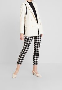 Blazer blanc double boutonnage, col roulé noir, pantalon slim à carreaux noir et blanc, et escarpins nude à bout pointu. Tissu texturé, design ajusté.