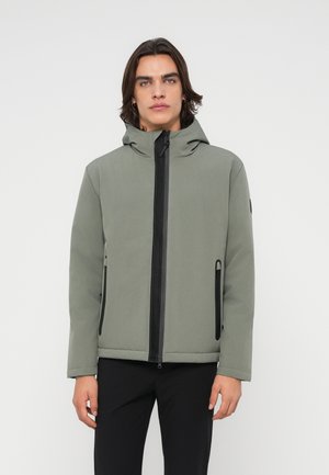 Jeune homme aux cheveux foncés mi-longs portant une veste à capuche gris clair avec une fermeture éclair noire et un pantalon noir, debout sur un fond blanc uni.