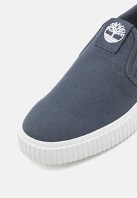 Scarpa slip-on in tela blu navy con suola in gomma bianca. Presenta una patch con logo con un design di albero nella parte superiore. Superficie in tessuto texturizzato.