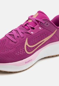 Paarse sportieve schoen met een mesh bovenkant, gouden Nike-logo en een gestructureerd patroon. Heeft roze accenten en een witte en lichtroze zool.
