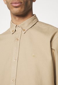 Camisa beige con cuello, cierres de botón, tejido ligero y un logo sutil en dorado. La textura es suave y tiene un corte clásico.