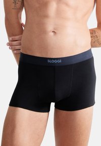 Schwarze Baumwoll-Boxershorts mit einem dunkelblauen Gummibund, der mit dem Text "SLOGGI" versehen ist. Das Design beinhaltet einen unterstützenden Frontbeutel und eine glatte Oberfläche.
