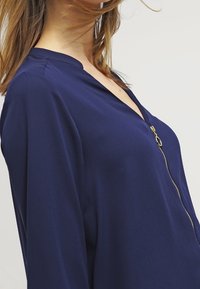 Námořnická modrá blůza se zipovým zapínáním, s výstřihem do V a dlouhými rukávy. Měkká textura a minimalistický design zdůrazňují jednoduchost.