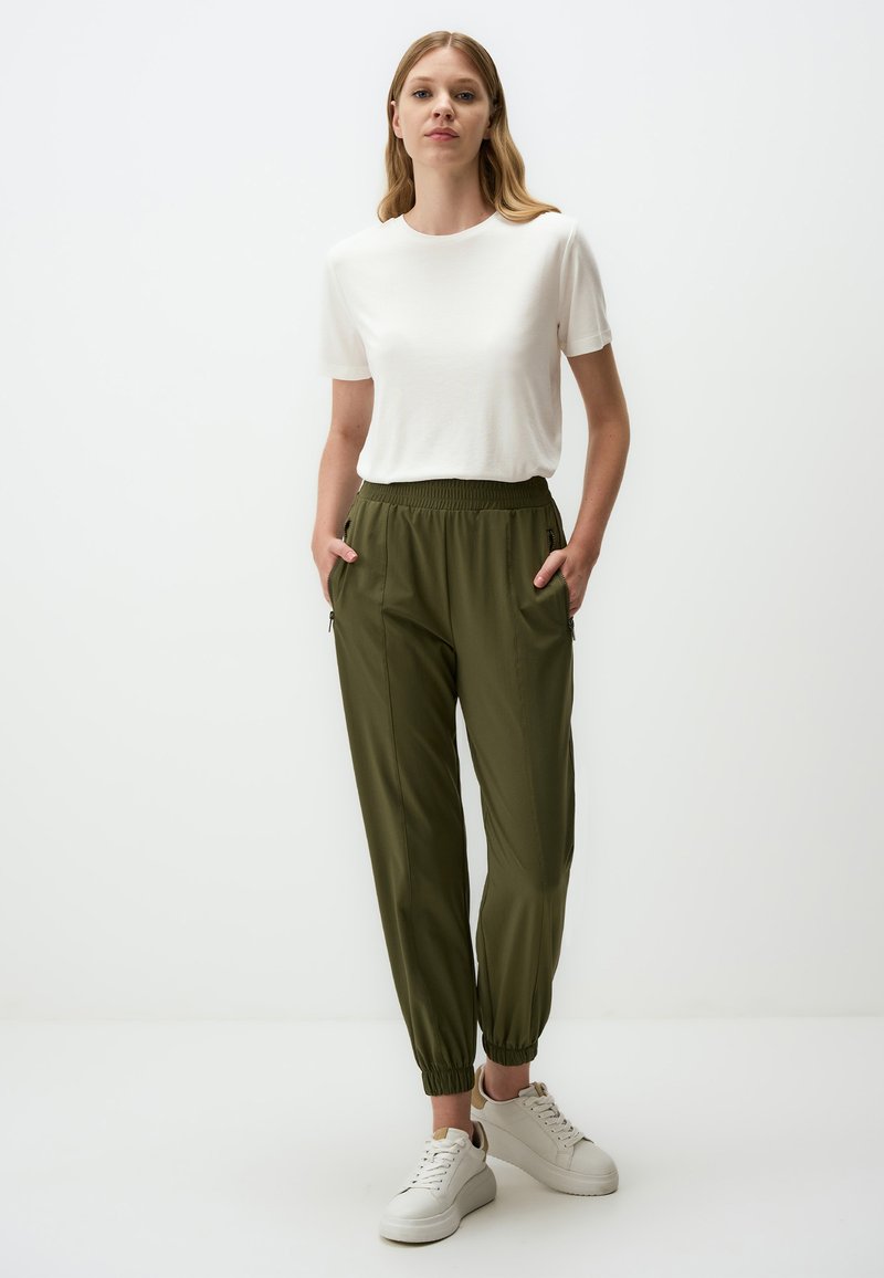 Witte korte mouwen shirt en olijfgroene rekbare broek met boorden, met twee zijzakken en een gladde stoftextuur. Witte sneakers.