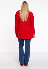 Rode tuniek met lange mouwen en een vloeiende uitstraling, gecombineerd met blauwe flare jeans en rode hakken. Soepele textuur, minimale details.