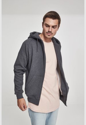 Junger Mann trägt einen geöffneten dunkelgrauen Kapuzenpullover über einem hellbeigen Shirt und hellblauen Jeans, blickt nach unten und zur Seite vor schlichtem Hintergrund.