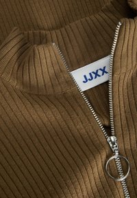 Maglione marrone a coste con colletto alto e zip argentata, dotato di un tirante circolare. L'etichetta riporta "JJXX" in testo blu su sfondo bianco.
