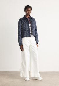 Marineblauwe gewatteerde jas met kraag, gecombineerd met een gestreept shirt en witte wijde broek. Model in witte sneakers staat op een neutrale ondergrond.