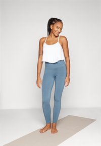 Weißes, kurzes Tanktop und blaue, hochgeschnittene Leggings aus dehnbarem Material, figurbetontes Design, stehend auf einer beige Yogamatte vor einem neutralen Hintergrund.