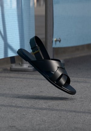 Μαύρο δερμάτινο σανδάλι με λουράκι αστραγάλου που αιωρείται πάνω από άσφαλτο κοντά σε μπλε κατασκευαστικό φράγμα.