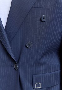Blazer croisé bleu marine à fines rayures avec des boutons noirs, porté sur un haut blanc, présentant une poche à rabat avec un détail boucle en argent.