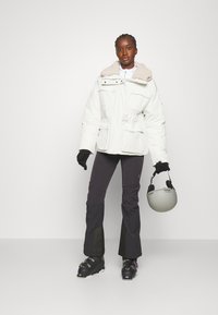 Witte pufferjack met een beige nepbont kraag, twee voorkruipen, zwarte handschoenen, zwarte skibroeken en een grijze helm in de hand.