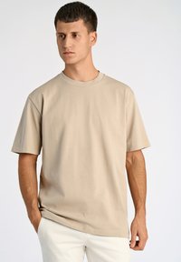Beige kortærmet T-shirt lavet af blødt stof, med en rund halsudskæring og en afslappet pasform, vist mod en ensfarvet baggrund.