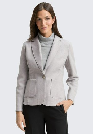 Frau trägt ein hellgraues, tailliertes Sakko über einem grauen Rollkragenpullover, mit schwarzen Hosen, steht vor einem schlichten hellen Hintergrund.