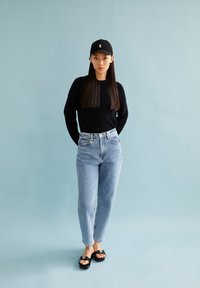 Pull en tricot noir, jean droit bleu clair, casquette noire avec logo et tongs noires. Tenue simple et décontractée devant un fond bleu.