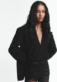 Blazer noir à coupe oversize, porté sur un haut à col en V profond et un short noir brillant à sequins. Textures lisses et formes contrastées tout au long.