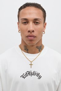 Biele tričko s čiernym textom "Ed Hardy", nosené so zlatým náhrdelníkom s krížovým príveskom a zlatou kruhovou náušnicou; tetovanie viditeľné na krku.