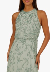 Haftowana sukienka bez rękawów w miętowym kolorze, zdobiona motywami kwiatów i liści z srebrnymi ozdobami, o warstwowej konstrukcji i miękkiej fakturze.